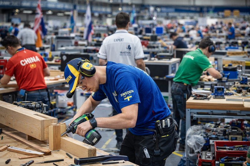 Tävlande i Yrkes-VM under internationell yrkestävling – unga deltagare utför yrkesmoment i en tävlingsarena, omgivna av verktyg, domare och internationella flaggor.